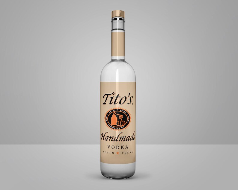 May include: A clear glass bottle of Tito's Handmade Vodka. The bottle has a tan label with black and orange text and a circular logo. The bottle is filled with a clear liquid and has a silver cap.