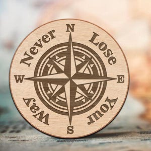 May include: A circular wooden compass rose with the words "Never Lose Your Way" engraved around the edge. The compass design features directional points and a central starburst pattern. The wood has a light brown colour.