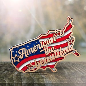 Op de afbeelding: Een houten uitsnede in de vorm van de Verenigde Staten, met het ontwerp van de Amerikaanse vlag. De woorden "American the beautiful" zijn in een cursief lettertype over elkaar geplaatst. De uitsnede is geschilderd in rood, wit en blauw.