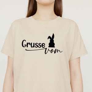 May include: A beige t-shirt featuring the text "Grusse vom" in black script, accompanied by a black silhouette of a rabbit. The shirt is a casual, short-sleeved style.