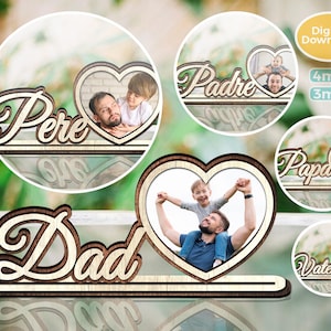 Può includere: Cornice portafoto in legno con la scritta "Dad" in un carattere decorativo, con un ritaglio a forma di cuore per una foto. Sono mostrate anche cornici aggiuntive con le parole "Pere", "Padre", "Papa" e "Vater".