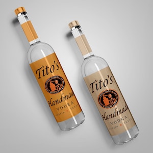 May include: Two clear glass bottles of Tito's Handmade Vodka. The bottles have gold-colored caps and labels with the brand name and logo in black and orange. The labels read "Handmade Vodka Austin Texas."