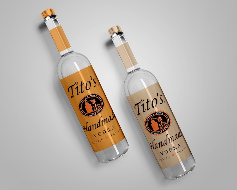 May include: Two clear glass bottles of Tito's Handmade Vodka. The bottles have gold-colored caps and labels with the brand name and logo in black and orange. The labels read "Handmade Vodka Austin Texas."