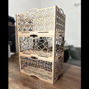 May include: A three-tiered wooden storage unit with intricate laser-cut designs. Each tier features a different pattern, including floral, geometric, and lattice styles. The unit has handles for easy carrying and is made of light-colored wood.
