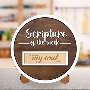 May include: A wooden, circular sign with the text "Scripture of the week" in white script. Below, a rectangular plaque reads "HE RESTORES my soul Psalm 23:3". The sign has a white border and a wooden stand.