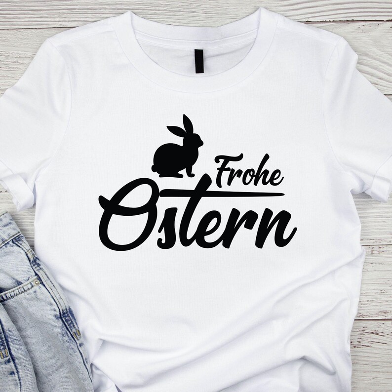 May include: White t-shirt featuring a black silhouette of a rabbit above the words "Frohe Ostern" in a stylish font. The design is centered on the shirt, with the text in a cursive style. The shirt is made of a soft material.