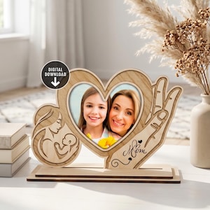 Può includere: Cornice in legno a forma di cuore, con una foto di madre e figlia. La cornice presenta un design intagliato di una madre e un bambino, e la parola "Mom" è scritta in corsivo. Si trova su una base di legno.