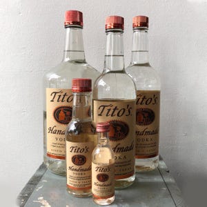 May include: Several clear glass bottles of Tito's Handmade Vodka with red caps. The bottles vary in size, from small sample bottles to large, with labels featuring the brand name and product details. The bottles are arranged on a light gray surface.