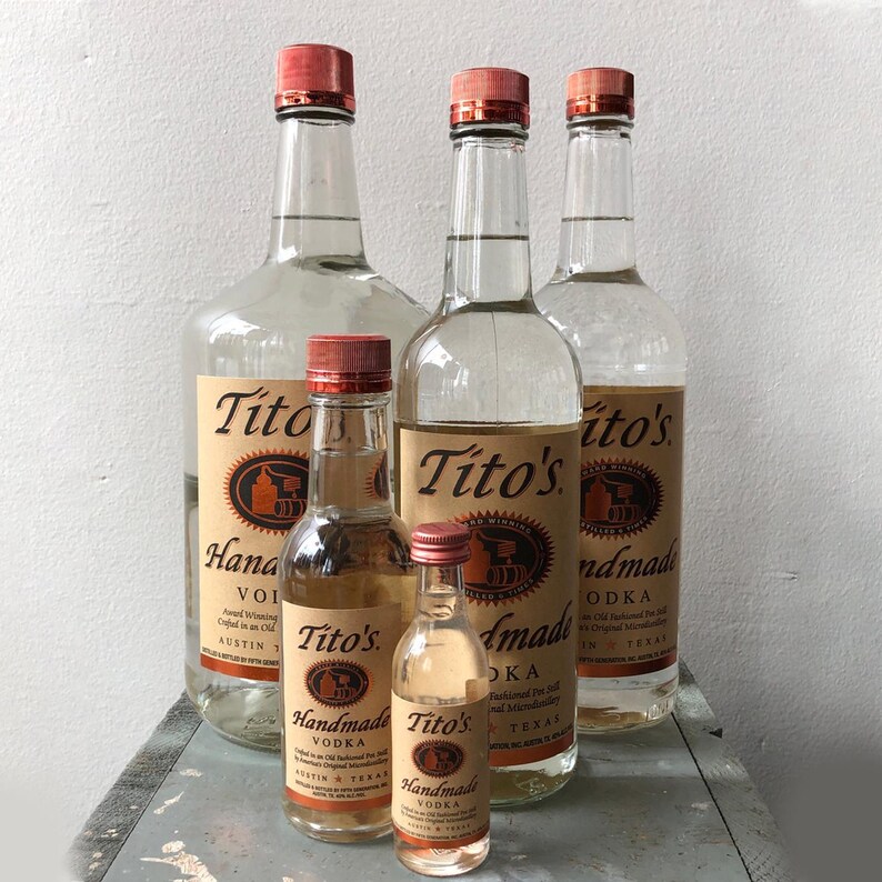 May include: Several clear glass bottles of Tito's Handmade Vodka with red caps. The bottles vary in size, from small sample bottles to large, with labels featuring the brand name and product details. The bottles are arranged on a light gray surface.
