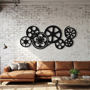 Könnte beinhalten: Schwarze Metall-Filmrollen-Wandkunst. Das Kunstwerk zeigt eine Ansammlung verschieden großer Filmrollen, die eine dynamische und künstlerische Darstellung erzeugen. Die Rollen sind an einer weißen Backsteinmauer angebracht.