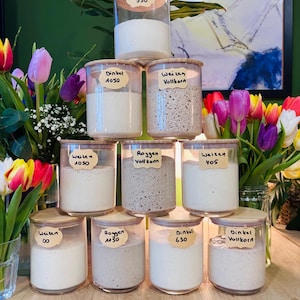 Könnte beinhalten: Eine Anordnung von Gläsern mit verschiedenen Mehlsorten, versehen mit handgeschriebenen Etiketten. Die Gläser sind auf einer Holzoberfläche gestapelt, mit frischen Tulpen im Hintergrund. Die Etiketten zeigen "Weizen 550", "Dinkel 1050" und "Weizen Vollkorn".