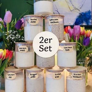 Könnte beinhalten: Eine Anordnung von Gläsern mit Holzdeckeln, jeweils beschriftet mit deutschem Text und Zahlen. Die Gläser enthalten verschiedene Mehlsorten, darunter "Weizen" und "Dinkel". Die Anordnung enthält ein "2er Set"-Etikett. Die Gläser stehen auf einer Holzoberfläche, mit bunten Blumen im Hintergrund.