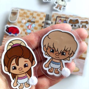 May include: Two cartoon stickers of children with light skin, brown and blonde hair, and large eyes. The girl has a bun and a pink and white diaper. The boy has a blue and white diaper. Additional stickers are visible in the background.