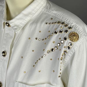 May include: A white denim shirt with a gold button and a design of gold stars and rhinestones on the shoulder.