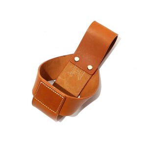 Puede incluir: Un soporte de cuero color canela para cinturón con un diseño curvo y costuras blancas visibles en los bordes. El accesorio incluye dos remaches metálicos de tono plateado que aseguran la correa superior al cuerpo principal, creando una estructura.