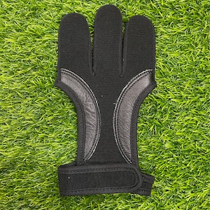 May include: Black archery glove with three finger stalls and a wrist strap. The glove features leather accents and is designed to protect the fingers during archery practice or competition. The glove is shown on a green surface.