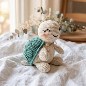 May include: A crocheted turtle toy with a cream-coloured body and a teal shell featuring a dark green stitched hexagonal pattern. The turtle has embroidered closed eyes, a small smile, pink cheeks, and a thin tan string tied around its neck.
