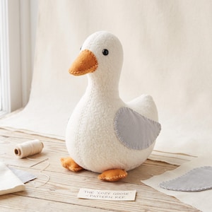 Puede incluir: Un juguete de ganso de peluche hecho de tela blanca texturizada con pico y patas naranjas, y alas de color gris claro unidas con costuras visibles. El ganso tiene pequeños ojos de cuentas negras. Una etiqueta de papel dice THE COZY GOOSE - PATTERN REF.