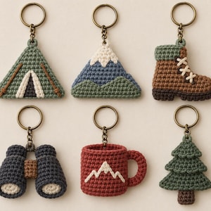 May include: A set of six crocheted keychains featuring outdoor-themed designs including a green tent, a blue and white mountain, a brown hiking boot, dark gray binoculars, a red mug with a mountain graphic, and a green pine tree with a brown trunk.