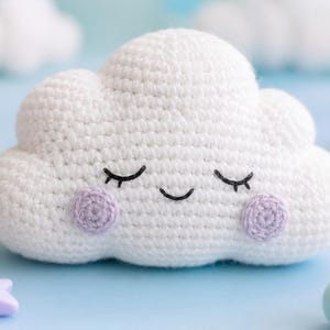 Puede incluir: Un juguete de peluche con forma de nube blanca tejida a ganchillo con una forma redondeada y esponjosa. La nube presenta ojos bordados con hilo negro con pestañas y una pequeña boca curva. Dos parches circulares de ganchillo morado claro.