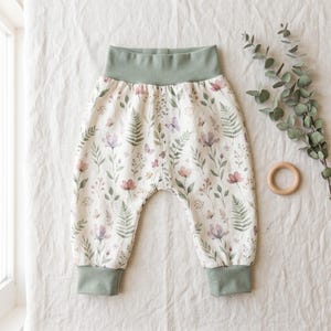 Op de afbeelding: Broek met elastische tailleband op een crèmekleurige basis met een botanische print van groene varens, kleine roze bloemen en vlinders. Het kledingstuk is voorzien van een brede, effen saliegroene tailleband en bijpassende geribbelde boorden.
