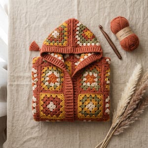 May include: A hooded cardigan made of crocheted granny squares in shades of burnt orange, mustard yellow, olive green, and cream. The garment features a ribbed front border, a hood with a small tassel, a ball of yarn, a wooden crochet hook, and dried grass.