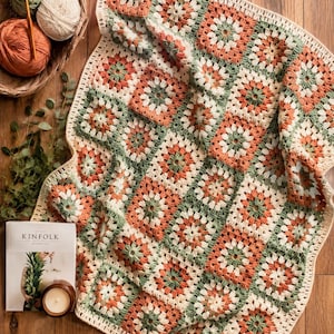 May include: A handmade crochet blanket featuring a repeating granny square pattern in shades of sage green, terracotta orange, and cream. The blanket has a scalloped cream border and is displayed alongside yarn balls and a Kinfolk magazine.