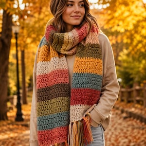 May include: A long, chunky crochet scarf featuring horizontal stripes in shades of mustard yellow, olive green, cream, dusty blue, and terracotta orange. The scarf is finished with thick, multi-coloured yarn tassels at both ends for a textured look.