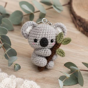 Può includere: Un piccolo portachiavi a forma di koala lavorato all'uncinetto in grigio, con testa rotonda, grandi orecchie con dettagli interni bianchi, occhi neri, naso nero e guance rosa. Il koala tiene un ramo lavorato all'uncinetto marrone con foglie verdi.