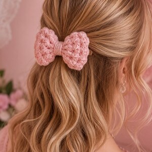 May include: A light pink crochet hair bow clip featuring a textured, knotted yarn design. The bow is constructed with a central wrapped yarn detail and is attached to a hair clip, securing a section of wavy blonde hair in a half-up style.