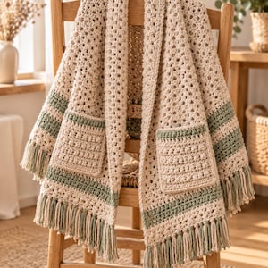 May include: A cream-colored crochet cardigan featuring a granny square pattern, two front pockets with a textured bobble stitch design, horizontal sage green stripes, and long fringe trim along the bottom hem and sleeve edges.