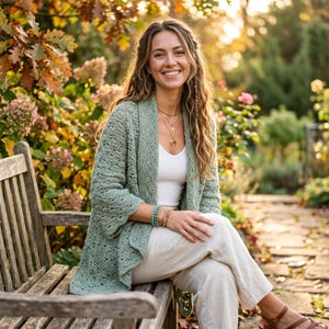 May include: A sage green open-front cardigan featuring an intricate crochet lace pattern with scalloped edges, worn over a white tank top and light-colored trousers, accessorized with layered necklaces and a stack of beaded bracelets on the wrist.