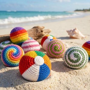 May include: A collection of round, crocheted yarn balls featuring various patterns including rainbow stripes, multicolored spirals, and segmented panels in red, white, blue, yellow, green, and pink, resting on a sandy surface.
