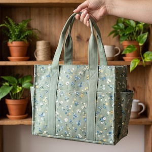 May include: A sage green canvas tote bag featuring a small floral pattern in white, blue, and yellow. The bag has two sturdy matching green fabric handles, a rectangular base, and exterior side pockets for storage, constructed with reinforced stitching.