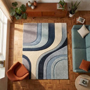 Könnte beinhalten: Ein rechteckiger Teppich mit Retro-Design. Der Teppich hat geschwungene Linien in Grau-, Blau- und Cremetönen. Der Teppich liegt auf einem Holzboden, und der Raum ist mit einem Sofa und einem Stuhl eingerichtet.
