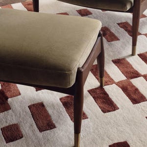 May include: A mid-century modern ottoman with a rectangular, olive-green upholstered seat. The wooden legs are dark brown with brass-colored tips. The ottoman sits on a cream-colored rug with a geometric pattern of brown rectangles.