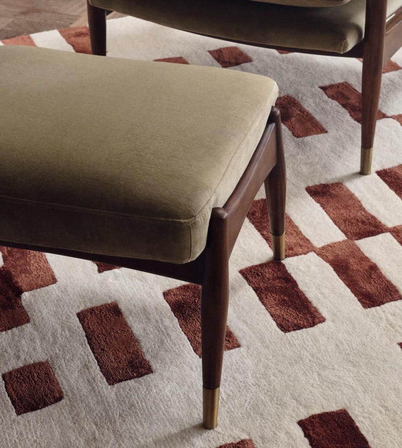 May include: A mid-century modern ottoman with a rectangular, olive-green upholstered seat. The wooden legs are dark brown with brass-colored tips. The ottoman sits on a cream-colored rug with a geometric pattern of brown rectangles.
