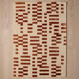 May include: Rectangular beige rug with a repeating pattern of rust-colored rectangular blocks. The blocks are arranged in a seemingly random, geometric design. The rug is made of a soft, textured material, suitable for a living room or bedroom.