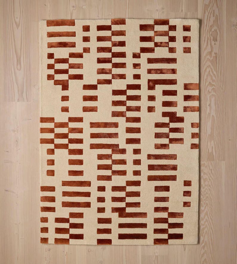 May include: Rectangular beige rug with a repeating pattern of rust-colored rectangular blocks. The blocks are arranged in a seemingly random, geometric design. The rug is made of a soft, textured material, suitable for a living room or bedroom.