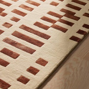 May include: A beige rug with a geometric pattern of brown rectangular shapes. The rug has a soft texture and is placed on a light-colored wooden floor. The design is modern and minimalist, suitable for various interior styles.