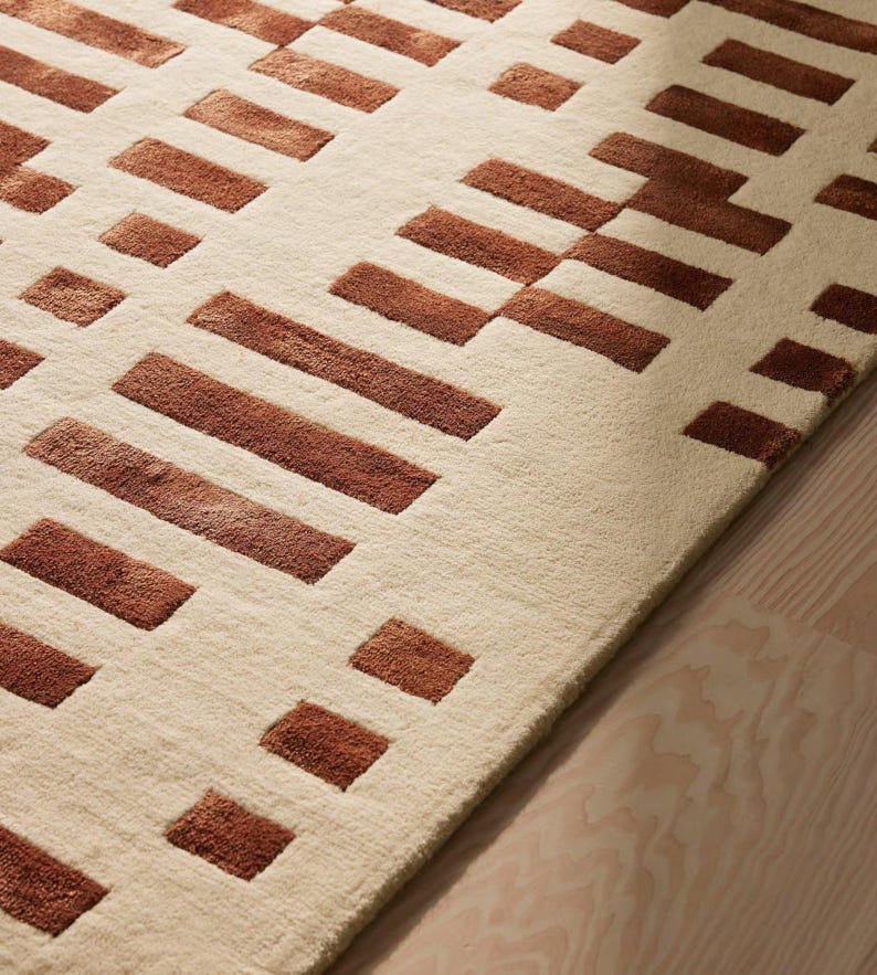May include: A beige rug with a geometric pattern of brown rectangular shapes. The rug has a soft texture and is placed on a light-colored wooden floor. The design is modern and minimalist, suitable for various interior styles.