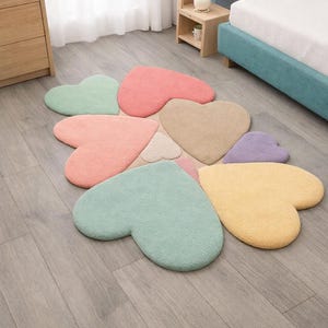 Könnte beinhalten: Eine Ansammlung herzförmiger Teppiche in Pastellfarben wie Rosa, Grün, Gelb und Beige, die auf einem Holzboden angeordnet sind. Die Teppiche sind weich und flauschig und schaffen eine verspielte Atmosphäre.