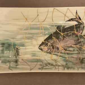 Könnte beinhalten: Rechteckige Servierplatte aus Keramik mit handgemaltem Fisch- und Netzdesign. Die Platte hat eine weiße Basis mit grünen und goldenen Zierleisten an den Griffen. Der Fisch ist in Grau- und Lilatönen dargestellt.