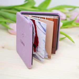 May include: A small, open, lavender-colored photo album with the name "EMMA" embossed on the cover. The album is made of a soft, plush material and contains several photographs. The pages are visible, showcasing various images.