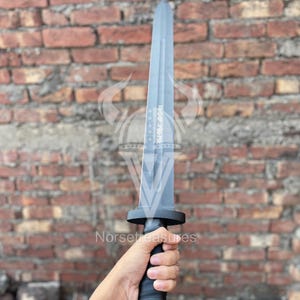 May include: A gray sword with a black handle and guard is held up against a brick wall. The sword has a long, flat blade and a textured handle. The text "Norse Treasures" is visible in the image.