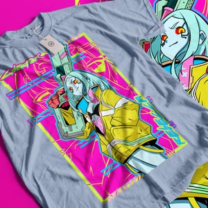 Op de afbeelding: Lichtblauw t-shirt met een opvallende afbeelding van een vrouwelijk personage met blauw haar, rode ogen en een gele jas, die een futuristisch wapen vasthoudt. Het ontwerp staat tegen een roze en gele geometrische achtergrond met het woord "Cyberpunk" in neon groen.