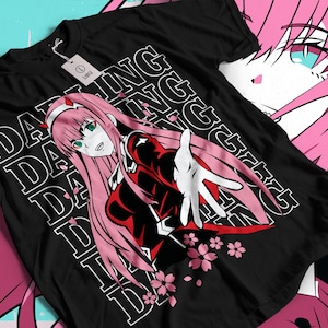 May include: Black t-shirt with a graphic of a pink-haired anime character with green eyes, reaching out. The design includes the text "DARLING" and "ING" in white, along with pink cherry blossom accents. A tag is attached to the shirt.