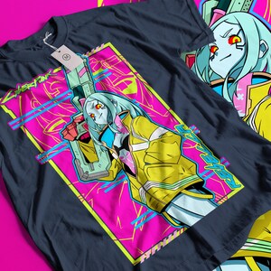 Op de afbeelding: Donker marineblauw T-shirt met een levendig grafisch ontwerp. Het ontwerp bevat een personage met blauw haar, een gele jas en een wapen, tegen een roze en gele achtergrond. Het woord "Cyberpunk" is zichtbaar in het ontwerp.