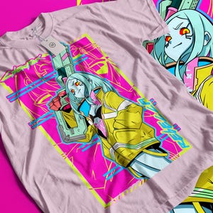 Op de afbeelding: Een lichtroze t-shirt met een levendig grafisch ontwerp. Het ontwerp toont een vrouwelijk anime-personage met blauw haar en een gele jas, die een futuristisch wapen vasthoudt. De achtergrond is een mix van roze, gele en blauwe geometrische vormen.