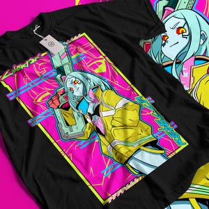 Op de afbeelding: Zwart t-shirt met een levendig grafisch ontwerp. Het ontwerp toont een personage met blauw haar, een gele jas en een wapen, tegen een roze en blauwe achtergrond. Het woord "CYBERPUNK" is ook zichtbaar.